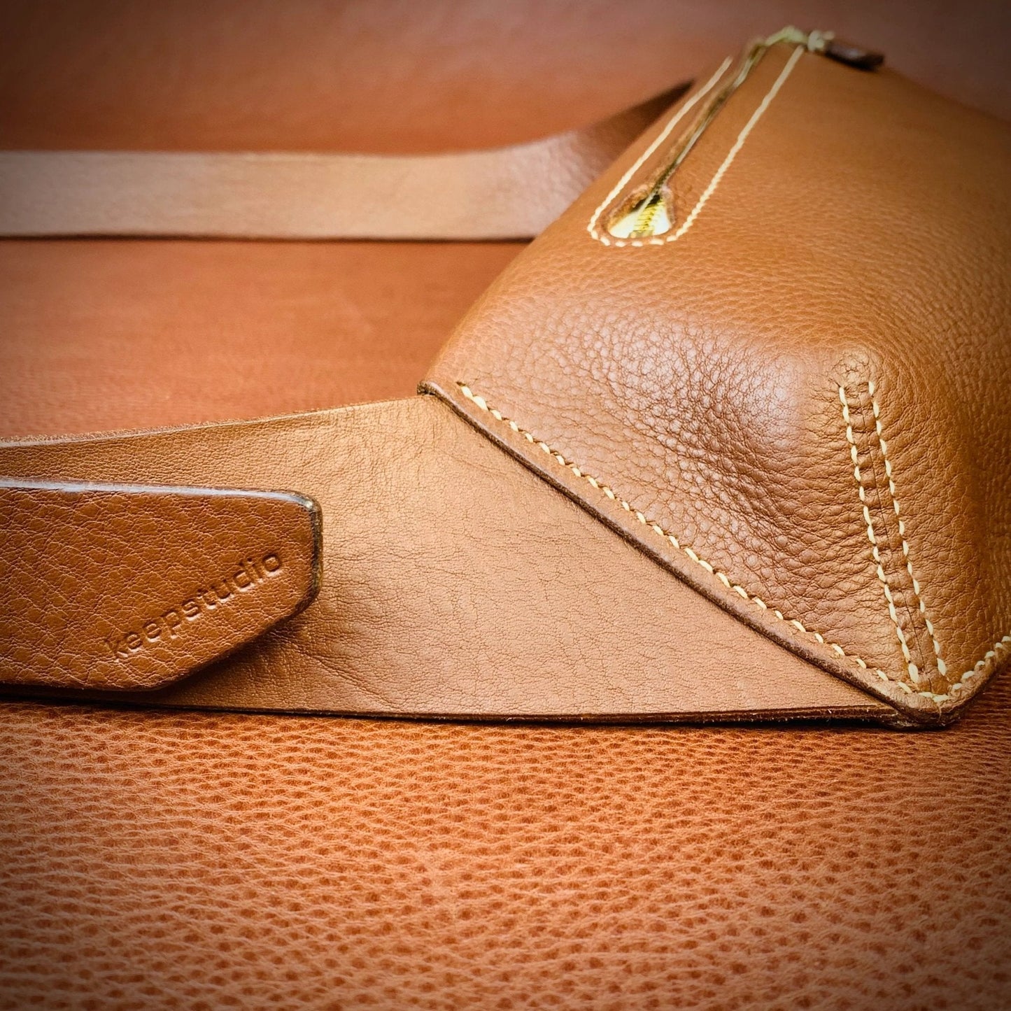 Close up detail of the hand stitching on a tan leather bag with a keep studio logo on the adjustable strap end
